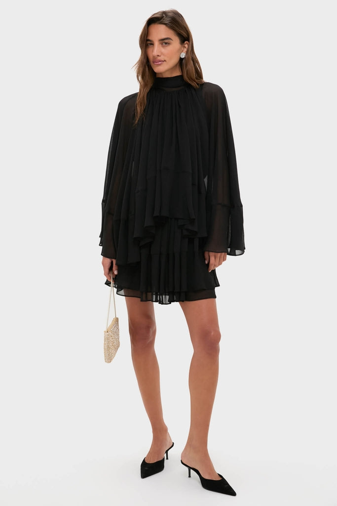 Model wearing the classic Black Cape Chiffon Mayfield Dress with layered ruffles, flared sleeves, paired with pointed-toe heels, and holding a chic embellished clutch for an effortless, high-end look.